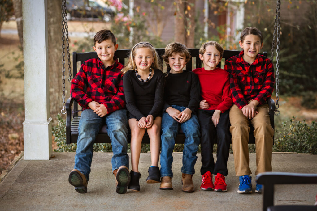 5 young children in red and black outfits are sitting on a porch swing at their home in Peachtree City, GA.
