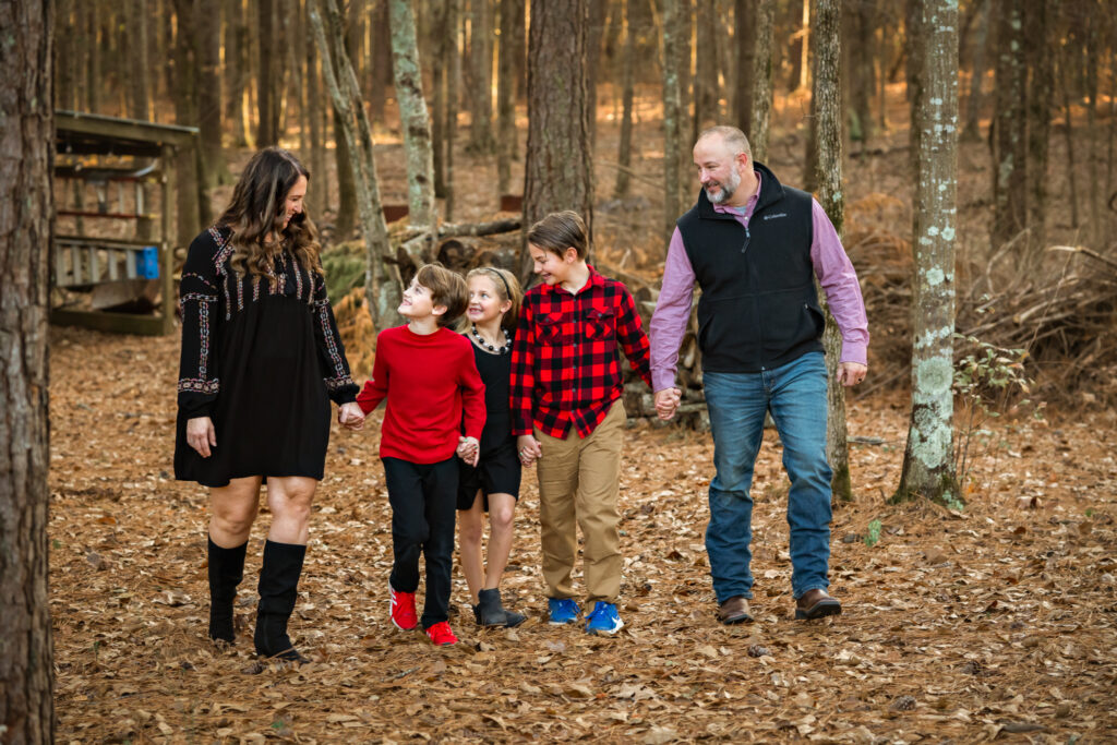 This family of 5 in Peachtree City are holding hands as they walk through a forest and smile at each other.