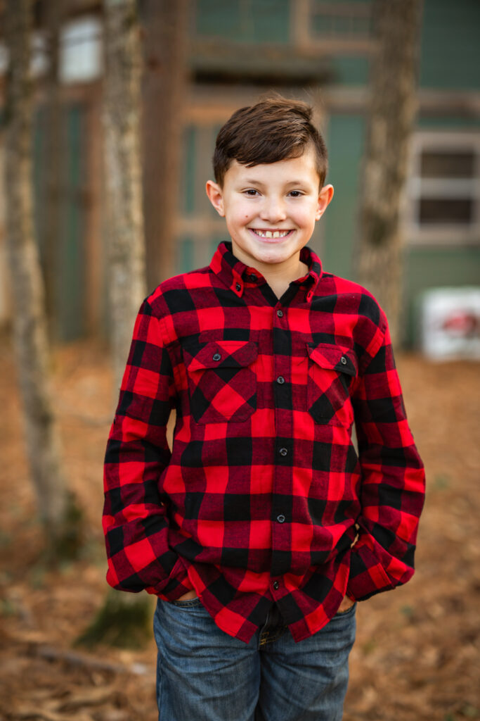 A young boy in red plaid has his hands in his pockets and is smiling for the camera at his family photography session.