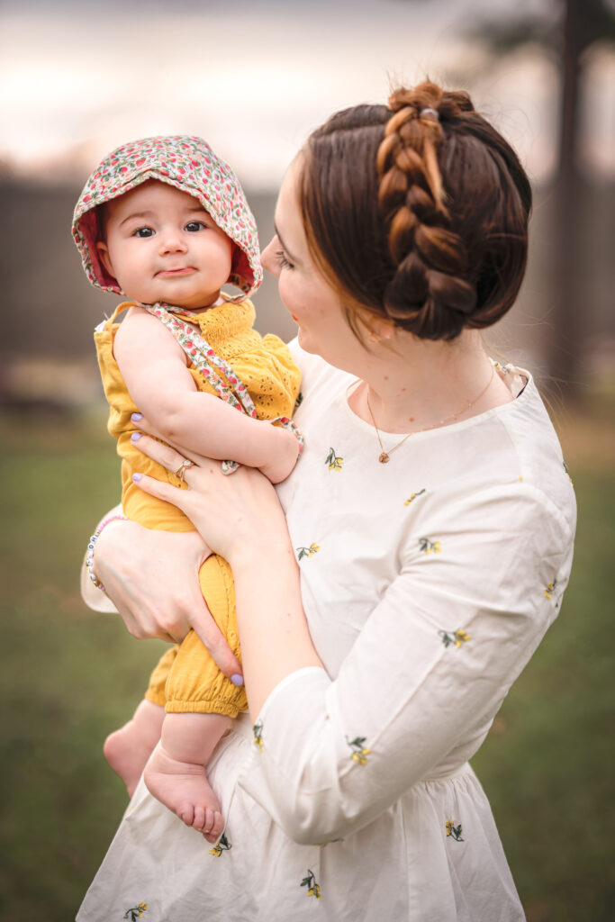 A mom in a white dress is looking at her young baby who is dressed in a yellow romper.