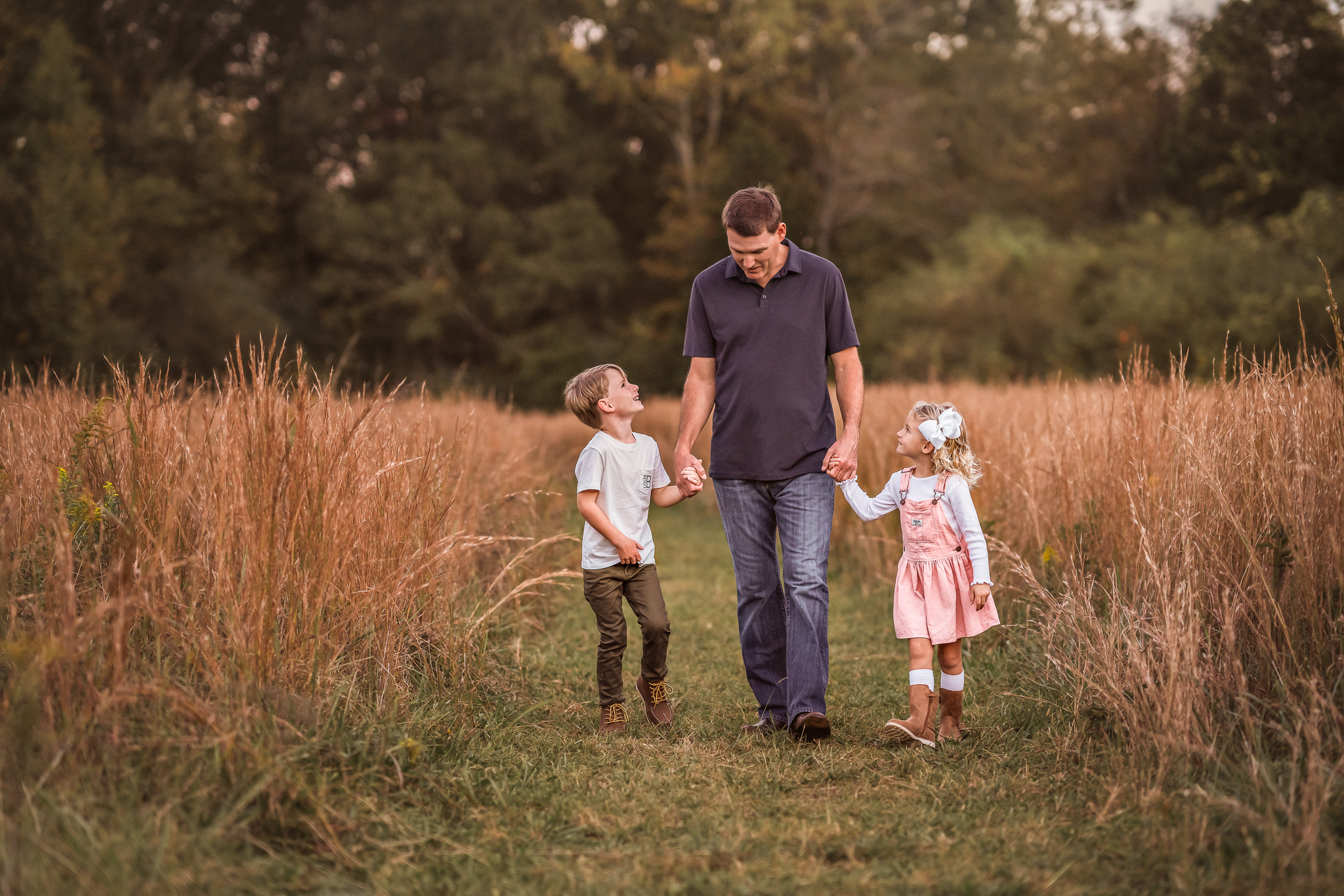 A dad is holding hands with his young children as they walk through a large field in Newnan, GA.