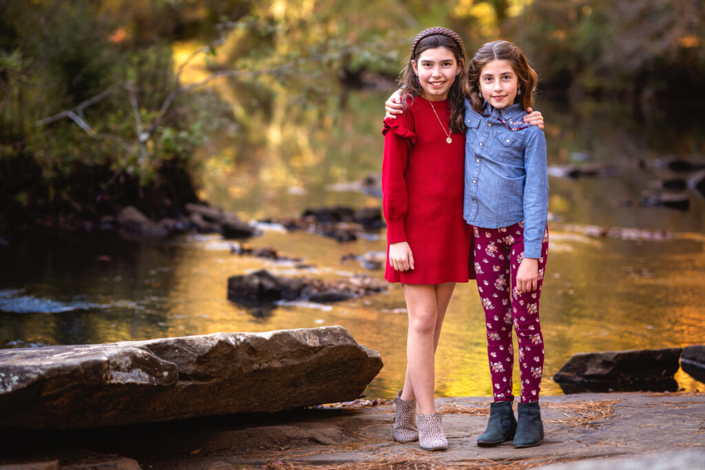 Two young sisters have their arms around each other as they smile for the camera in front of a stream.