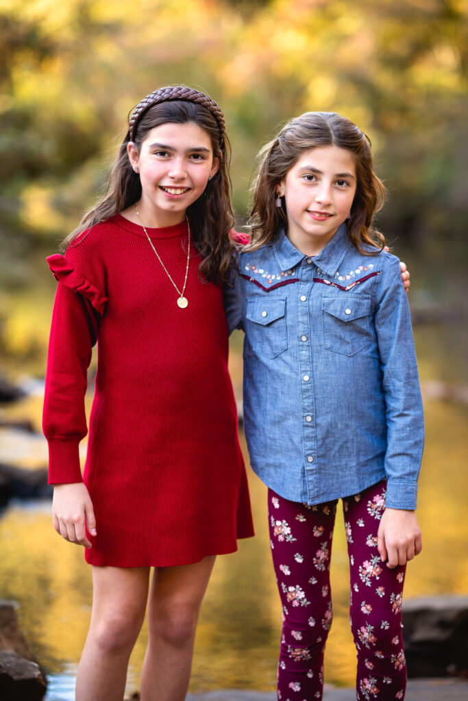 Two young sisters have their arms around each other and they are both smiling for family pictures in front of a small stream.