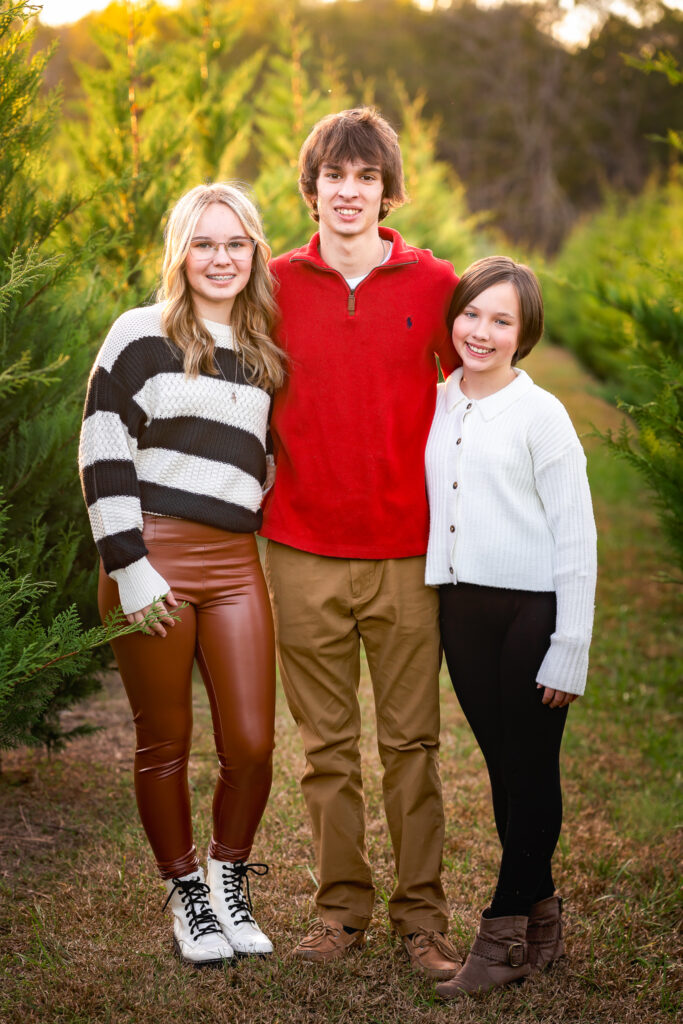Three young siblings embrace and smile as they stand in front of the pine trees at Minter's Farm. 
