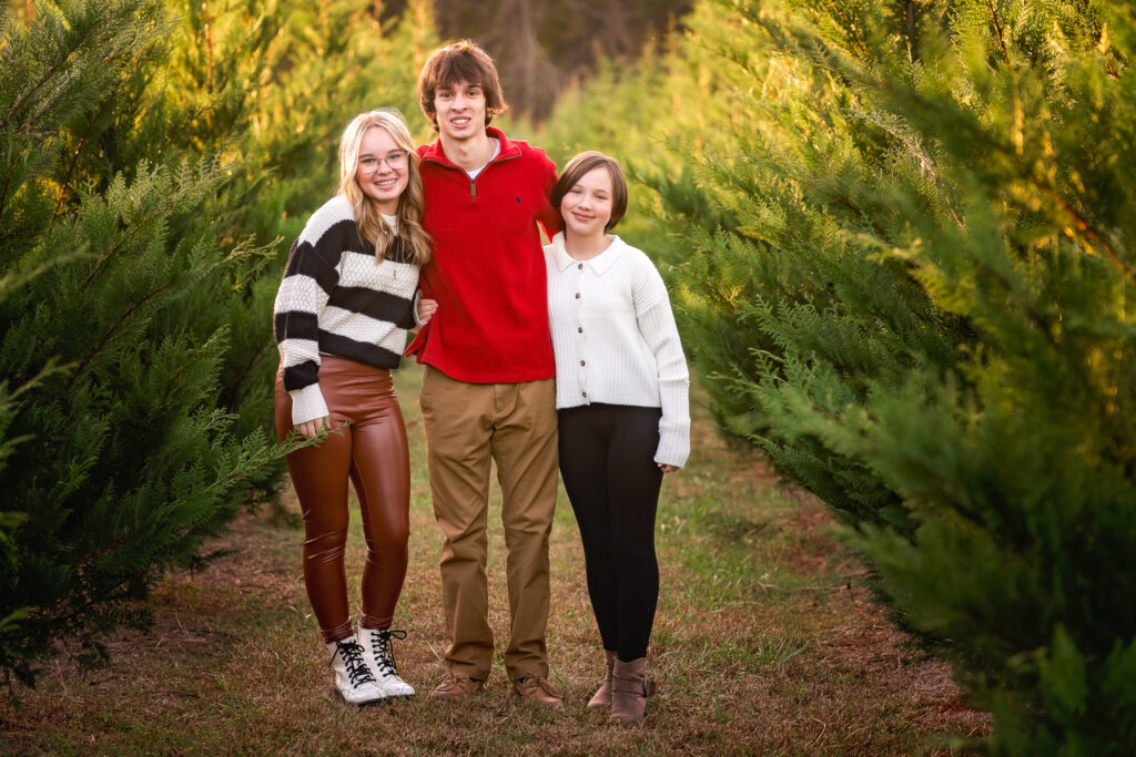 Three siblings dressed in sweaters are smiling for the camera as they take family pictures with Ashley Zohil Photography.