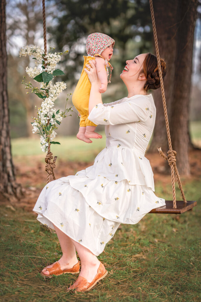 A mom in a white dress is sitting in a wooden swing and holding up her young baby who is dressed in yellow. 