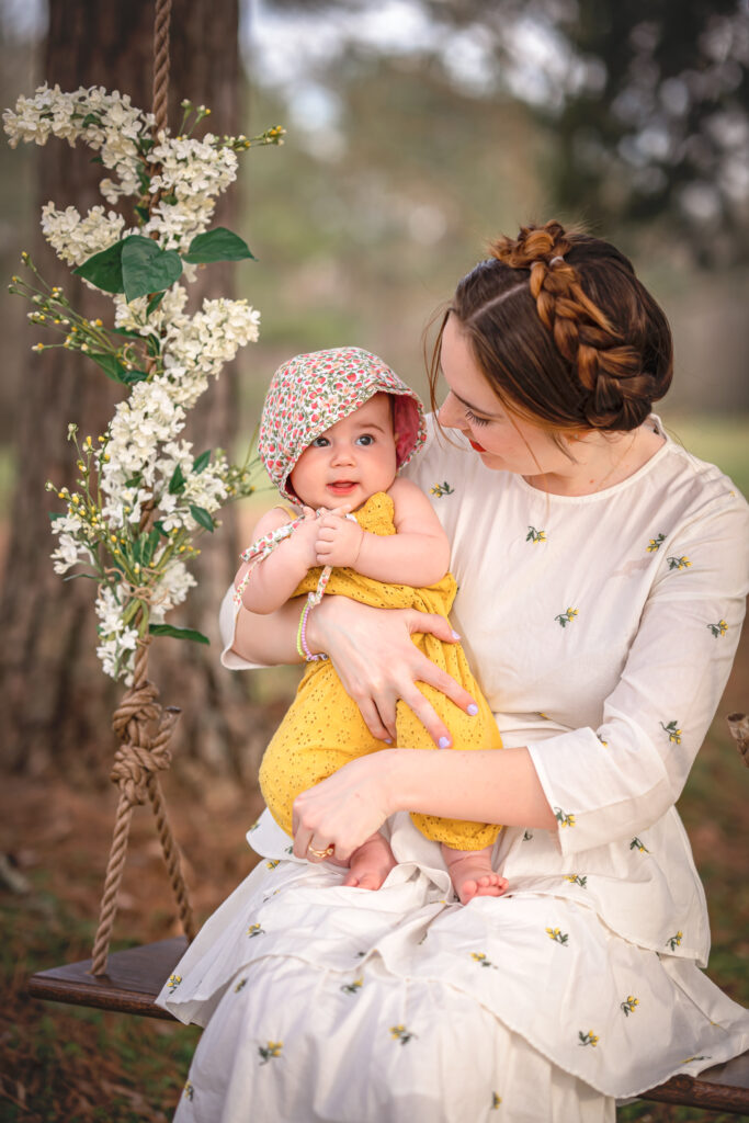 A mom in a white floral dress is sitting in a wooden swing and she is looking at the daughter she's holding. 
