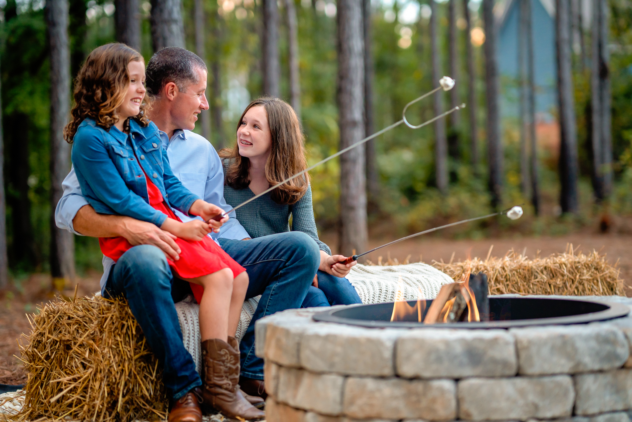 A dad is sitting with his two daughters as they roast marshmallows over a fire.
