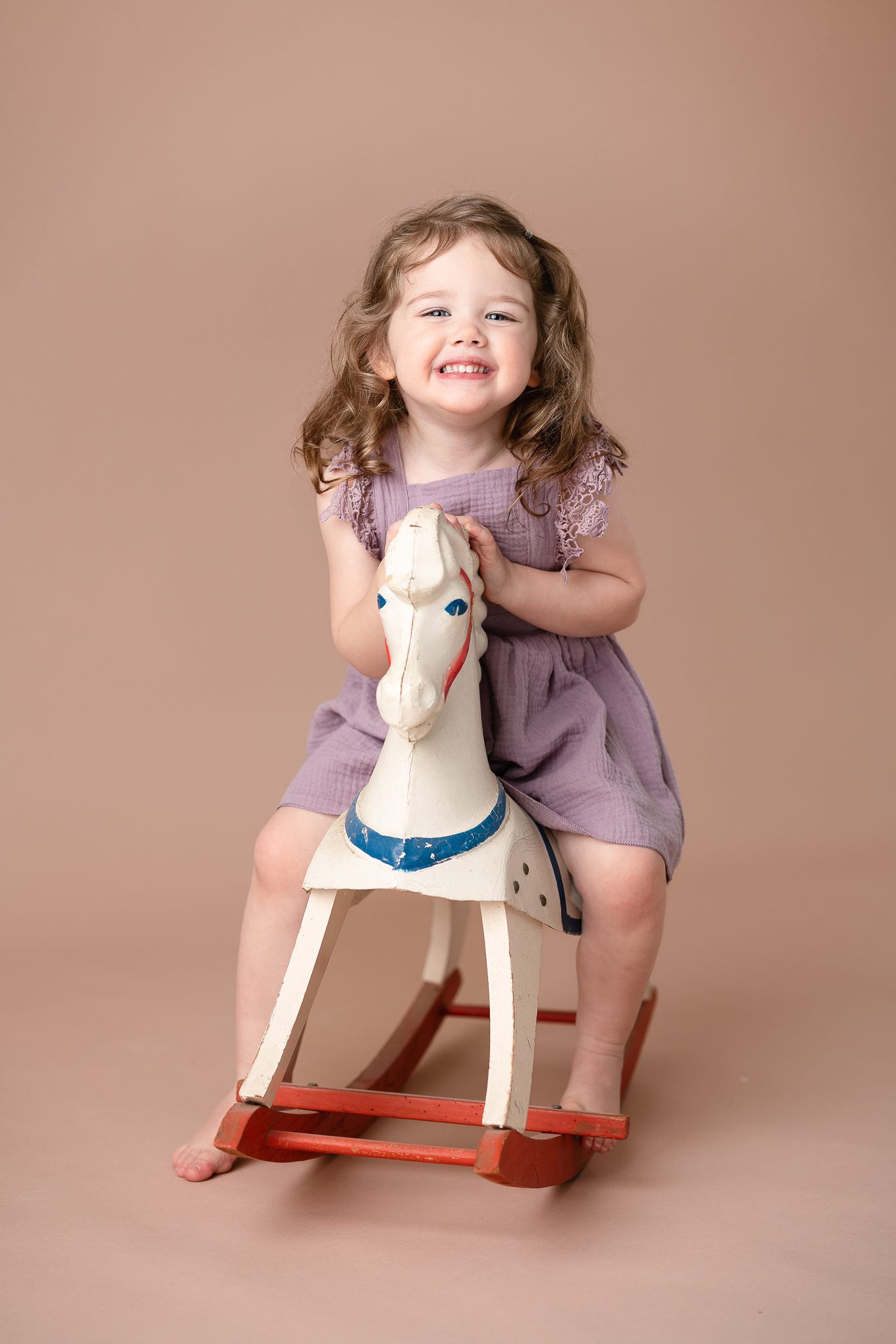 A little girl in a purple dress is smiling as she sits on a colorful wooden rocking horse.