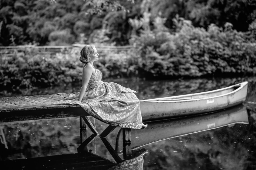 A black and white portrait of a high school senior girl sitting on the dock and looking out onto the water at Turnipseed Nursery Farms. 