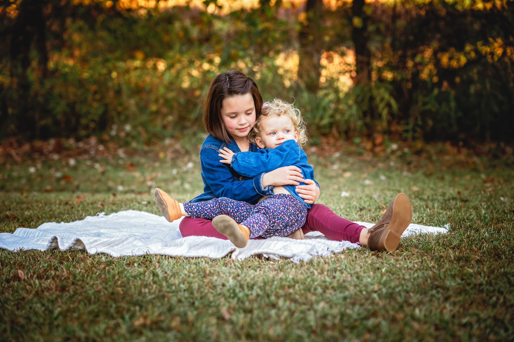 Two young sisters are sitting in a field and cuddling each other.